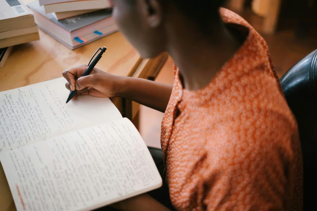 Focused black woman writing in a notebook with stacked books on a desk, illustrating learning.