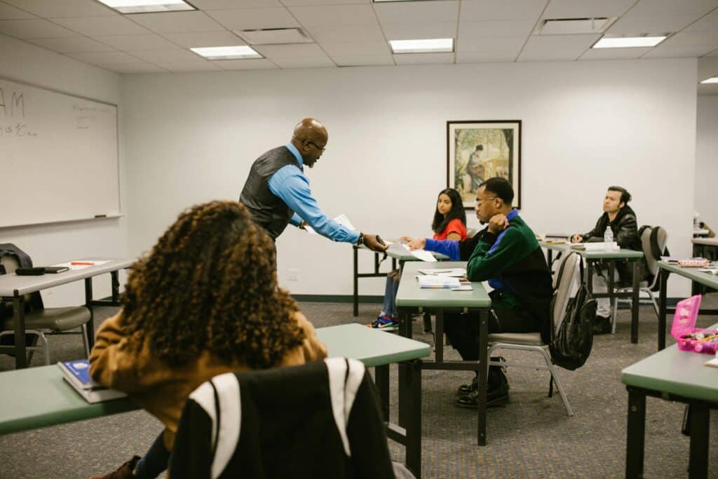 College classroom scene with diverse students and a teacher distributing papers, promoting education.
