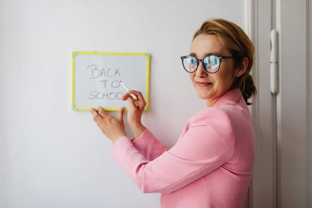 A cheerful female teacher in a pink blazer writes 'Back to School' on a whiteboard.