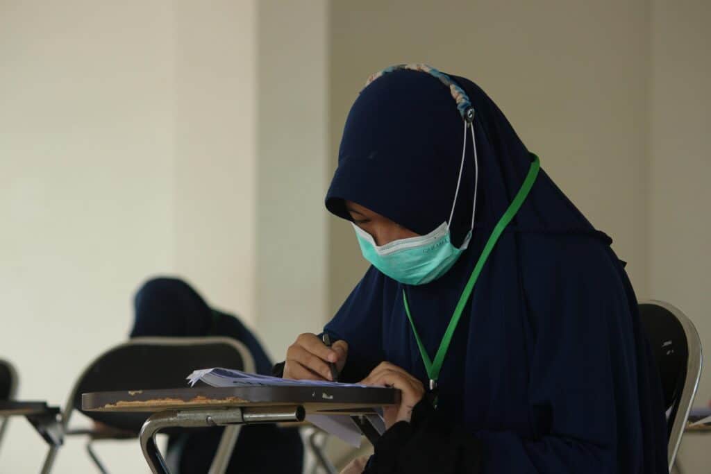 A Muslim woman in a hijab and face mask writing on paper during an exam indoors.